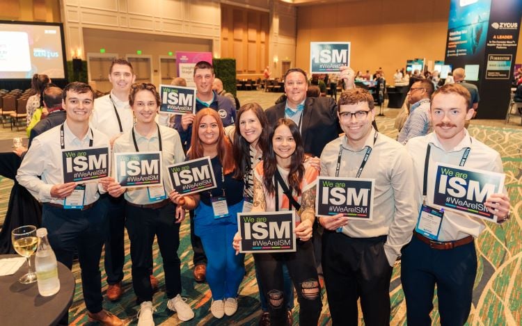 A group of young professionals stand together holding ISM signs in a large conference room