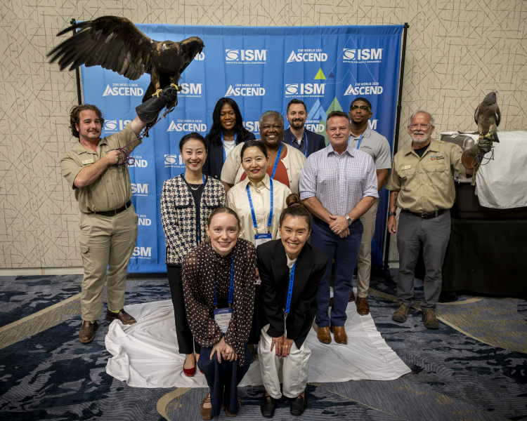 A group of professionals stand in front of an ISM World 2026 backdrop surrounded by two falcon handlers and falcons